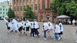 Grupa dwudziestu pięciu uczniów klasy piątej w pelerynach typu krzyżackiego idzie kolumną za przewodnikiem, zwiedzając Toruń.