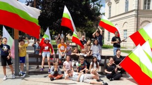 Zdjęcie zbiorowe - Na zdjęciu stoi grupa dzieci, niektórzy trzymają w rękach duże flagi w kolorach Lublina (biało-zielono-czerwony) oraz prowadzący warsztaty żonglerki flagami.