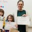 chłopiec i dziewczynka stoją z nauczycielką pod tablicą i trzymają dyplomy i nagrody z międzyszkolnego konkursu Przytulanki na dobranoc